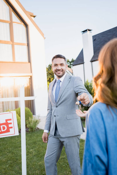 Smiling broker looking at camera while giving keys to blurred woman on foreground