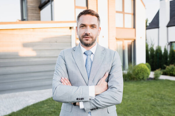 Уверенный брокер со скрещенными руками смотрит в камеру с размытыми домами на заднем плане