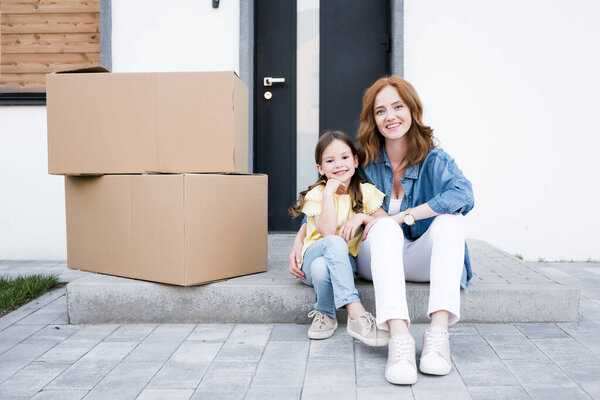 Smiling redhead mother hugging daughter while sitting on doorstep near carton boxes