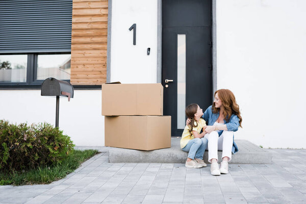 Happy mother and daughter looking at each other while sitting near carton boxes on doorstep