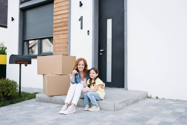 Happy redhead woman looking at key and hugging daughter while sitting on doorstep near cardboard boxes