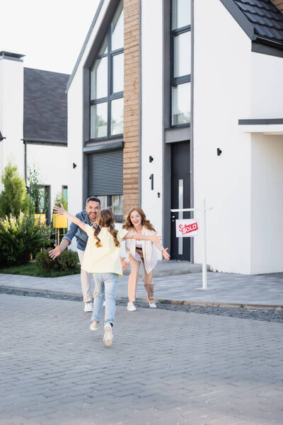 Back view of daughter with open arms running to happy parents near house and sign with sold lettering