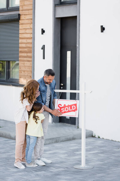 Full length of family hugging while looking at sign with sold lettering near house