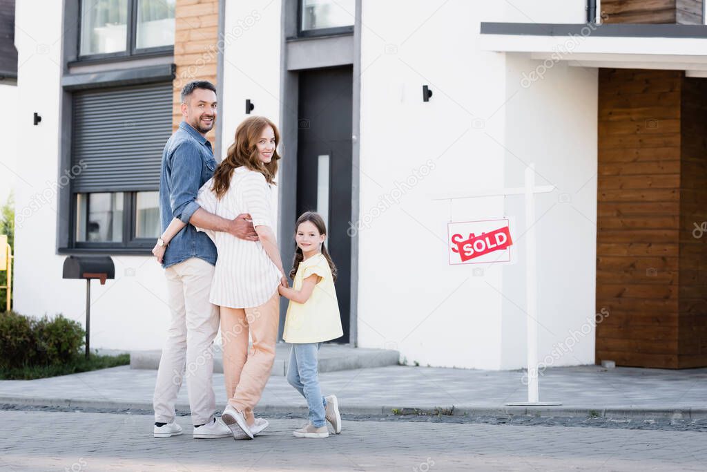 Full length of happy family with daughter looking at camera while standing near house and sign with sold lettering