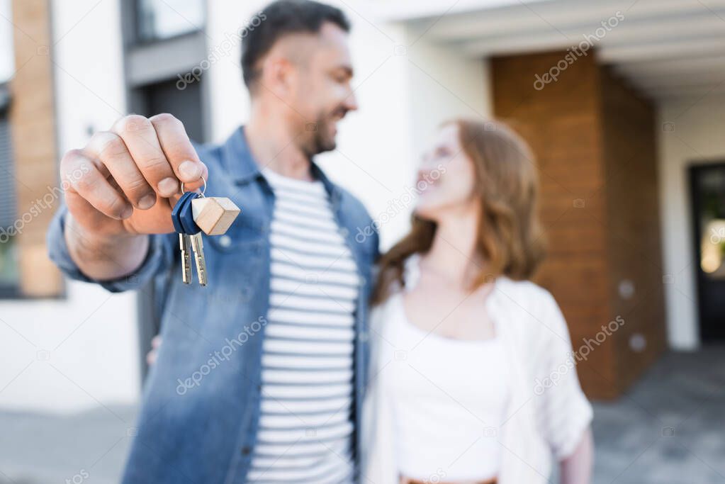 Keys and fob in hands of happy husband looking and hugging wife near house on blurred background