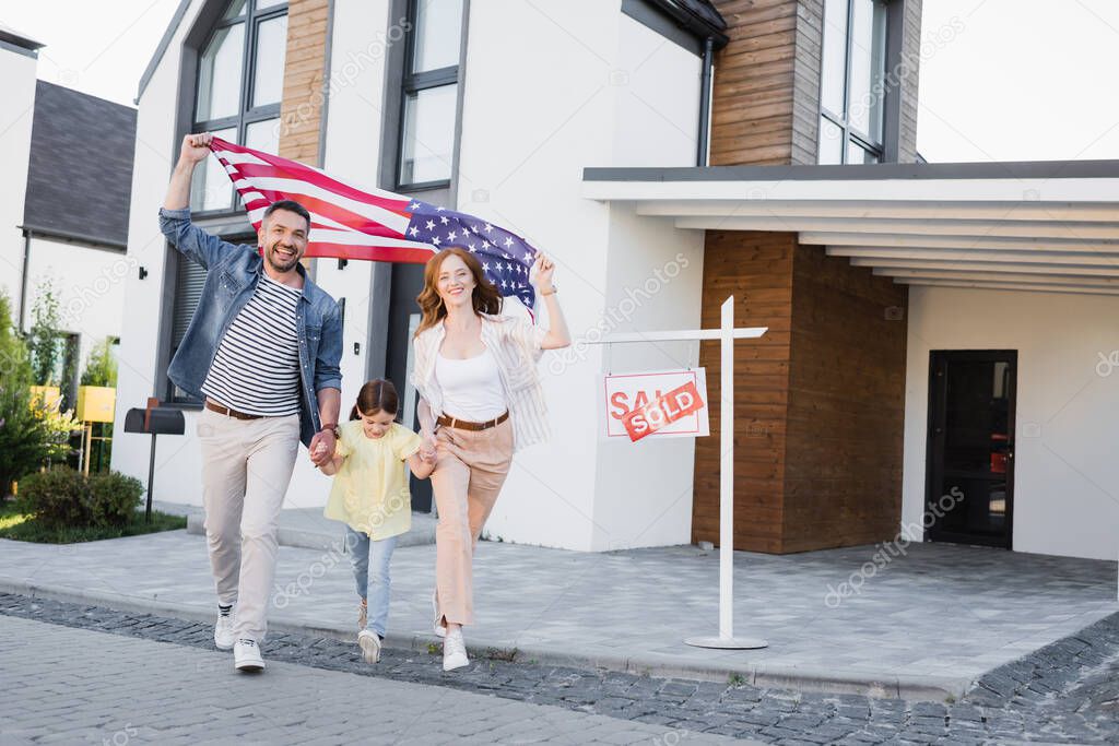 Daughter with happy mom and dad holding american flag going forward near sign with sold lettering and house