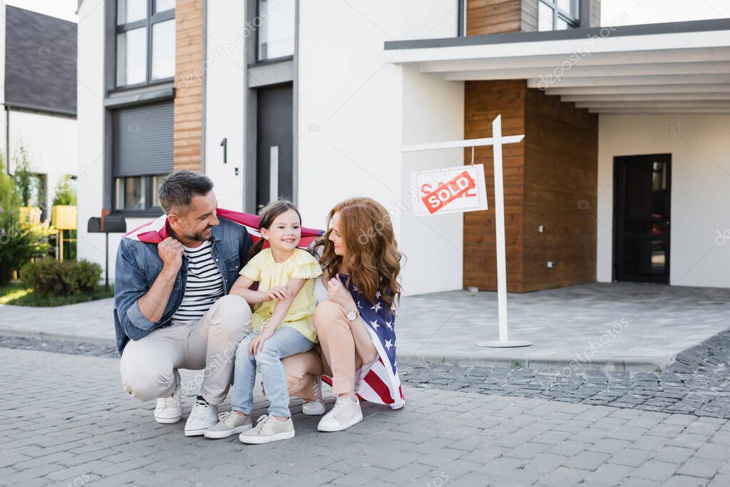 Happy couple with american flag looking at daughter while squatting near sign with sold lettering and house