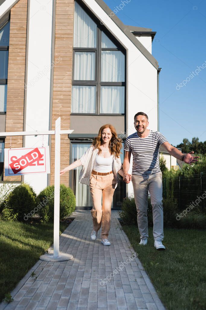Cheerful couple holding hands while running forward near sign with sold lettering and modern house