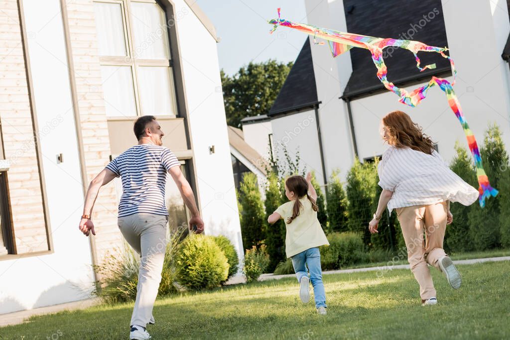 Back view of family running while flying kite on lawn near home