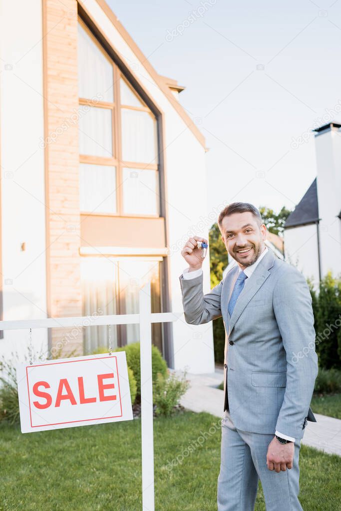 Happy broker with key looking at camera while standing near sign with sale lettering