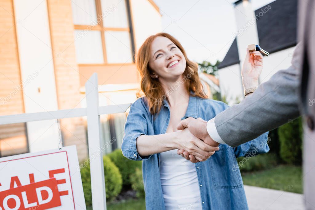 Happy redhead woman with keys shaking hands with broker near sign with blurred houses on background