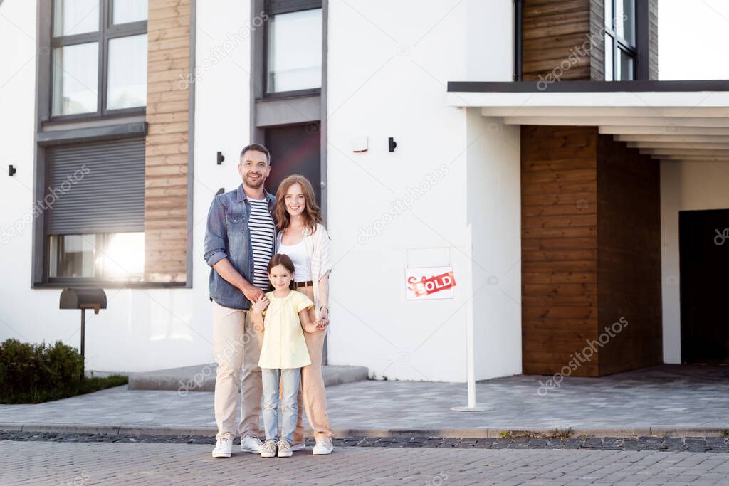 Full length of happy family hugging and looking at camera near house and sign with sold lettering