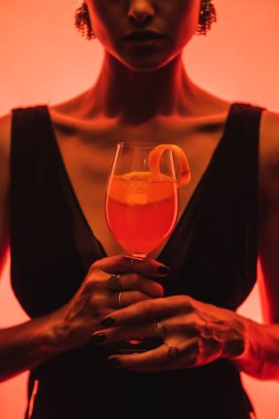 cropped view of elegant woman holding glass with mixed cocktail and orange on pink  clipart
