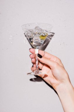 Beyaz üzerine alkol damlalarının yanında zeytinli bir kadeh martini sallayan kadın manzarası.
