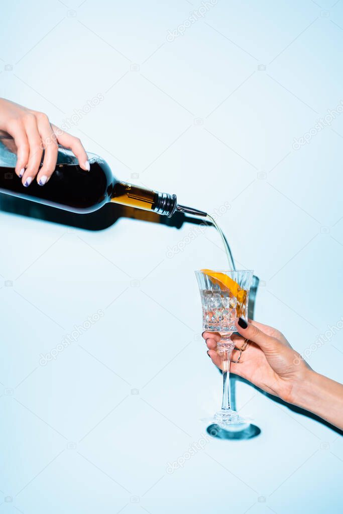 cropped view of woman holding bottle and pouring alcohol drink in glass with sliced orange on blue