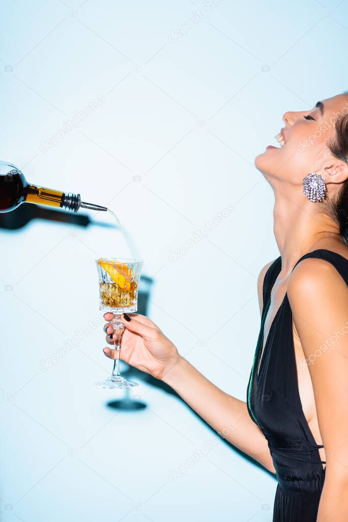 side view of happy woman laughing while holding alcohol drink in glass with sliced orange near bottle on blue