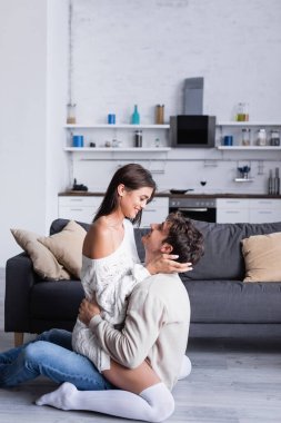 Gülümseyen adamın oturma odasında esmer kız arkadaşına sarılışının yan görüntüsü. 