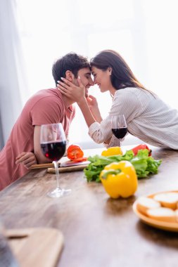 Gülümseyen kadın ön planda şarap ve sebzelerin yanında sevgilisinin yüzüne dokunuyor. 