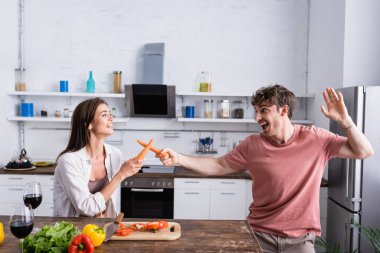 Cheerful couple fighting with carrots near vegetables and wine  clipart