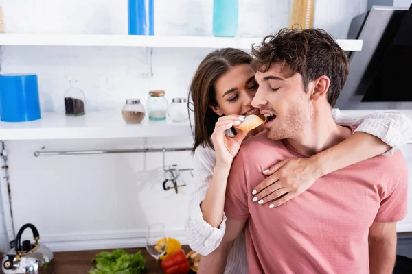 Smiling Woman Feeding Boyfriend Baguette Vegetables Blurred Background Kitchen — Stock Photo, Image