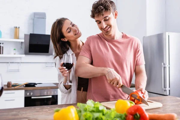 一个微笑的女人 在男朋友旁边拿着酒杯 在厨房里切红辣椒 — 图库照片