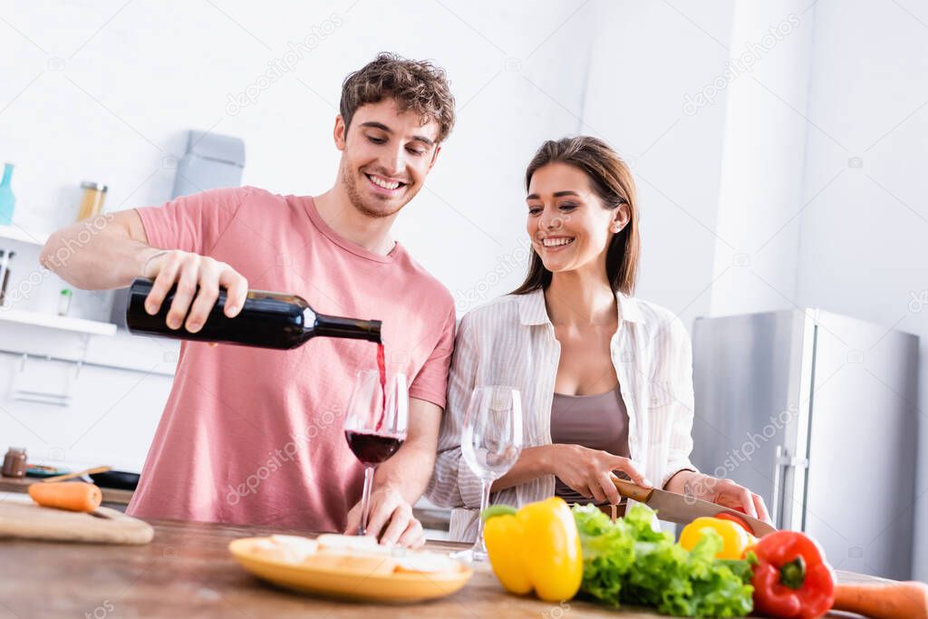 Smiling man pouring wine near girlfriend with knife and vegetables on blurred foreground 