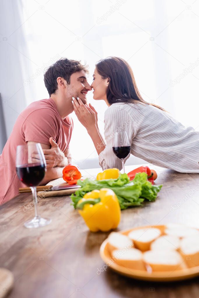 Smiling woman kissing boyfriend near vegetables and glasses of wine on blurred foreground 