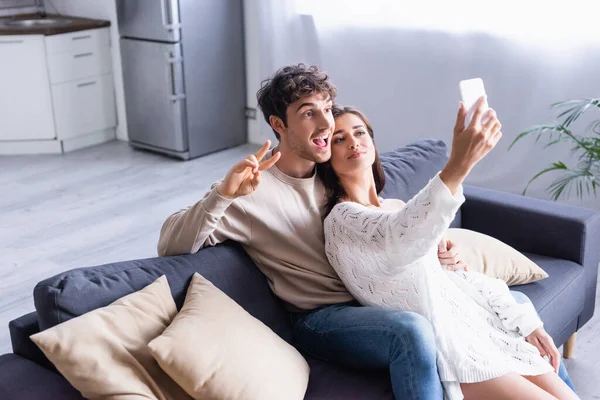 Cheerful Man Showing Peace Sign Girlfriend Taking Selfie Smartphone Couch — Stock Photo, Image