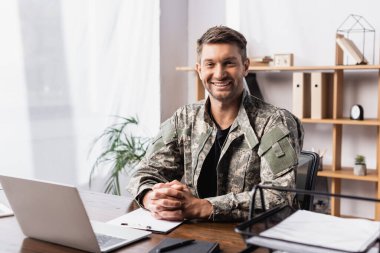 Üniformalı neşeli asker elleri dizüstü bilgisayarın yanında oturuyor.
