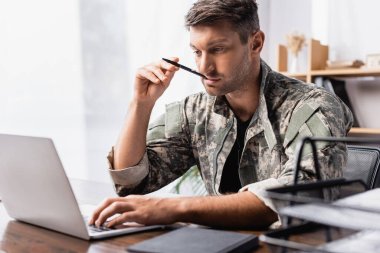 pensive soldier in uniform holding pen and using laptop near document tray on blurred foreground clipart