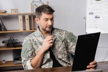 soldier in military uniform holding pen and looking at clipboard  clipart