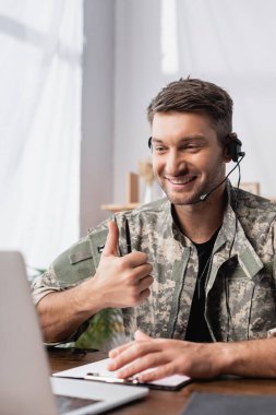 Askeri üniformalı ve kulaklıklı bir adam başparmağını dizüstü bilgisayarın yanında gösteriyor.