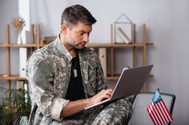 Amerikan bayrağı yanında dizüstü bilgisayar kullanan vatansever bir asker. 