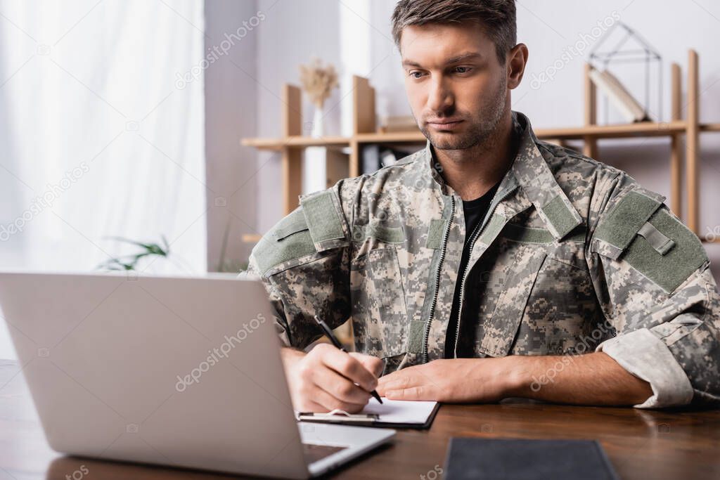 military man in uniform holding pen while writing on clipboard near laptop on desk