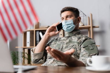 Tıbbi maskeli asker Amerikan bayrağı yakınındaki akıllı telefondan konuşuyor bulanık ön planda