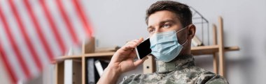 Tıbbi maskeli asker Amerikan bayrağı yakınındaki akıllı telefondan konuşuyor bulanık ön planda, afişte