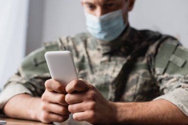 Akıllı telefon üniformalı askerin elinde ve tıbbi maske bulanık arka planda.