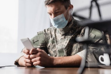Akıllı telefon üniformalı askerin elinde ve tıbbi maske ön planda bulanık bir belgesel tepsisinin yanında.