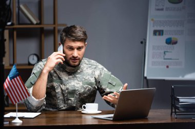 Üniformalı vatansever asker Amerikan bayrağı, fincanı ve dizüstü bilgisayarının yanında akıllı telefondan konuşuyor. 