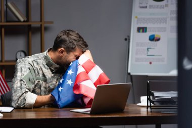 Üniformalı asker ağlıyor ve dizüstü bilgisayarın yanında Amerika bayrağıyla yüzünü örtüyor. 