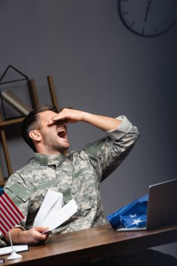 üniformalı kızgın asker gözlerini kapatıyor ve dizüstü bilgisayarın ve Amerikan bayraklarının yanında bağırırken elinde bir mektup tutuyor.
