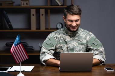 Amerikan bayrağı ve aletlerinin yanında dizüstü bilgisayar kullanan üniformalı mutlu asker. 