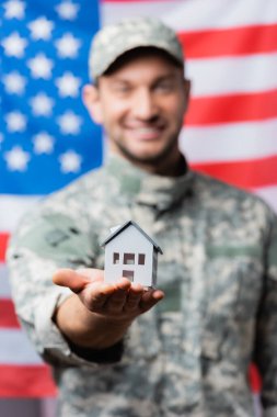 Bulanık arka planda Amerikan bayrağı yakınında üniformalı mutlu bir askerin ev modeli.