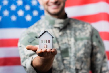 Bulanık arka planda Amerikan bayrağı yakınında üniformalı mutlu bir askerin ev modeli.