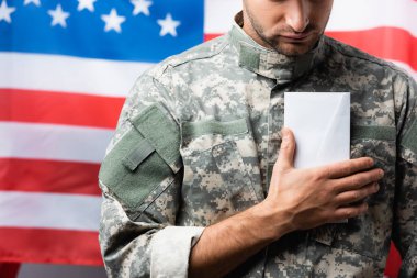 Amerikan bayrağının yakınındaki bulanık arkaplanda üniformalı vatansever asker görüntüsü.