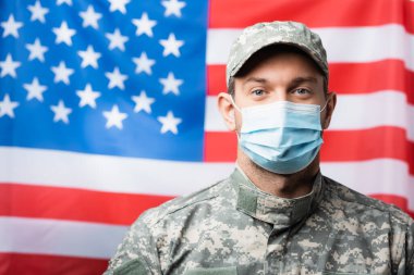 Üniformalı ve sağlık maskeli vatansever bir asker bulanık arka planda Amerikan bayrağı taşıyor.