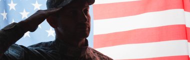 Üniformalı ve şapkalı vatansever asker arka planda Amerikan bayrağının yanında selam veriyor.