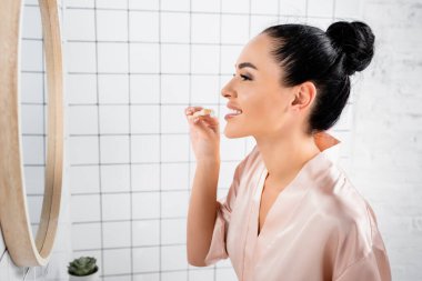 İpek bornozlu gülümseyen kadın banyoda diş fırçasıyla macun tutuyor. 