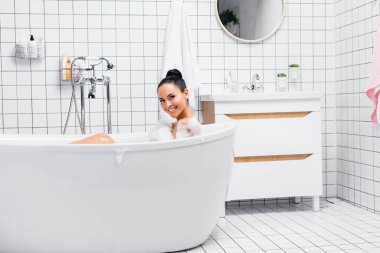 Neşeli esmer kadın köpükle banyo yaparken kameraya bakıyor. 