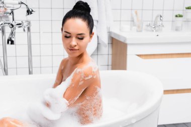 Esmer kadın evde köpükle banyo yaparken ıslak omzuna bakıyor. 